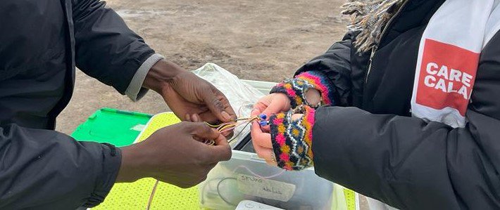 A person in a Care4Calais vest holds out their arm. Another person is pulling on threads to create a friendship bracelet for them.